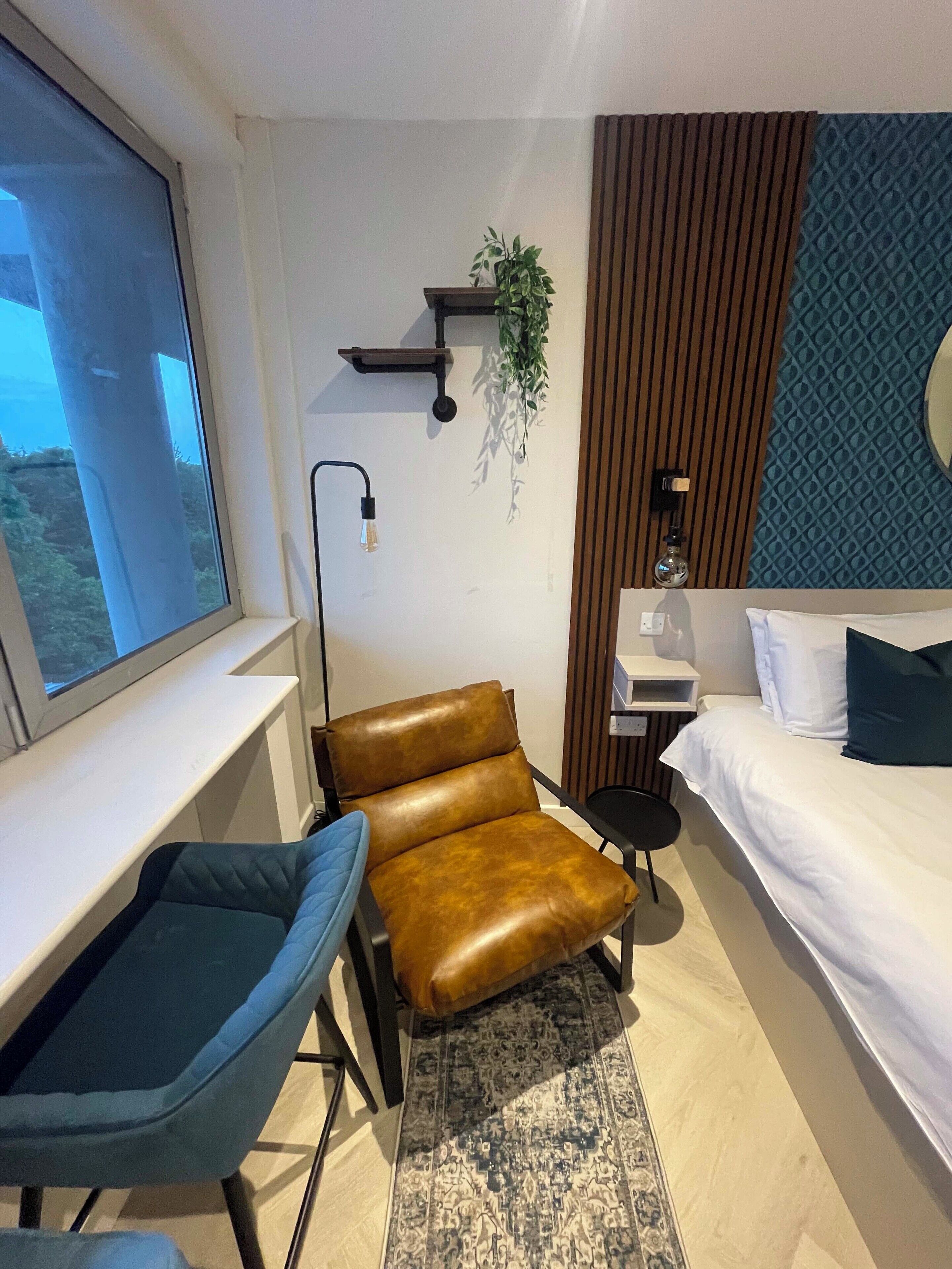 A modern hotel room with a large window, a brown leather chair, a small desk, a bed with white and dark pillows, a patterned rug, and a wall-mounted plant.