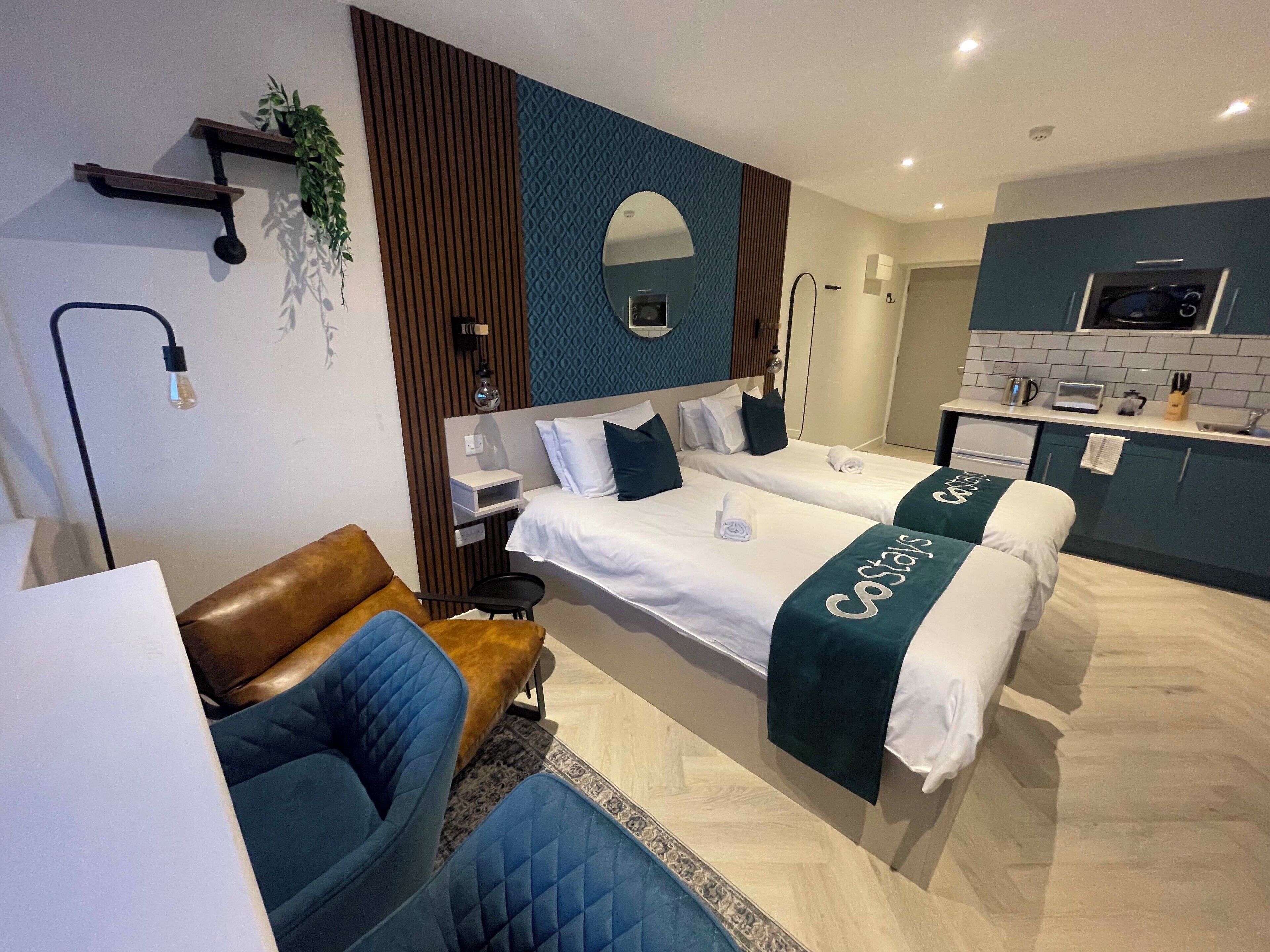 A modern hotel room with two beds, a seating area, a small kitchenette, and a bathroom.