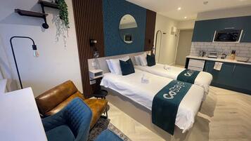 A modern hotel room with two beds, a seating area, a small kitchenette, and a bathroom.
