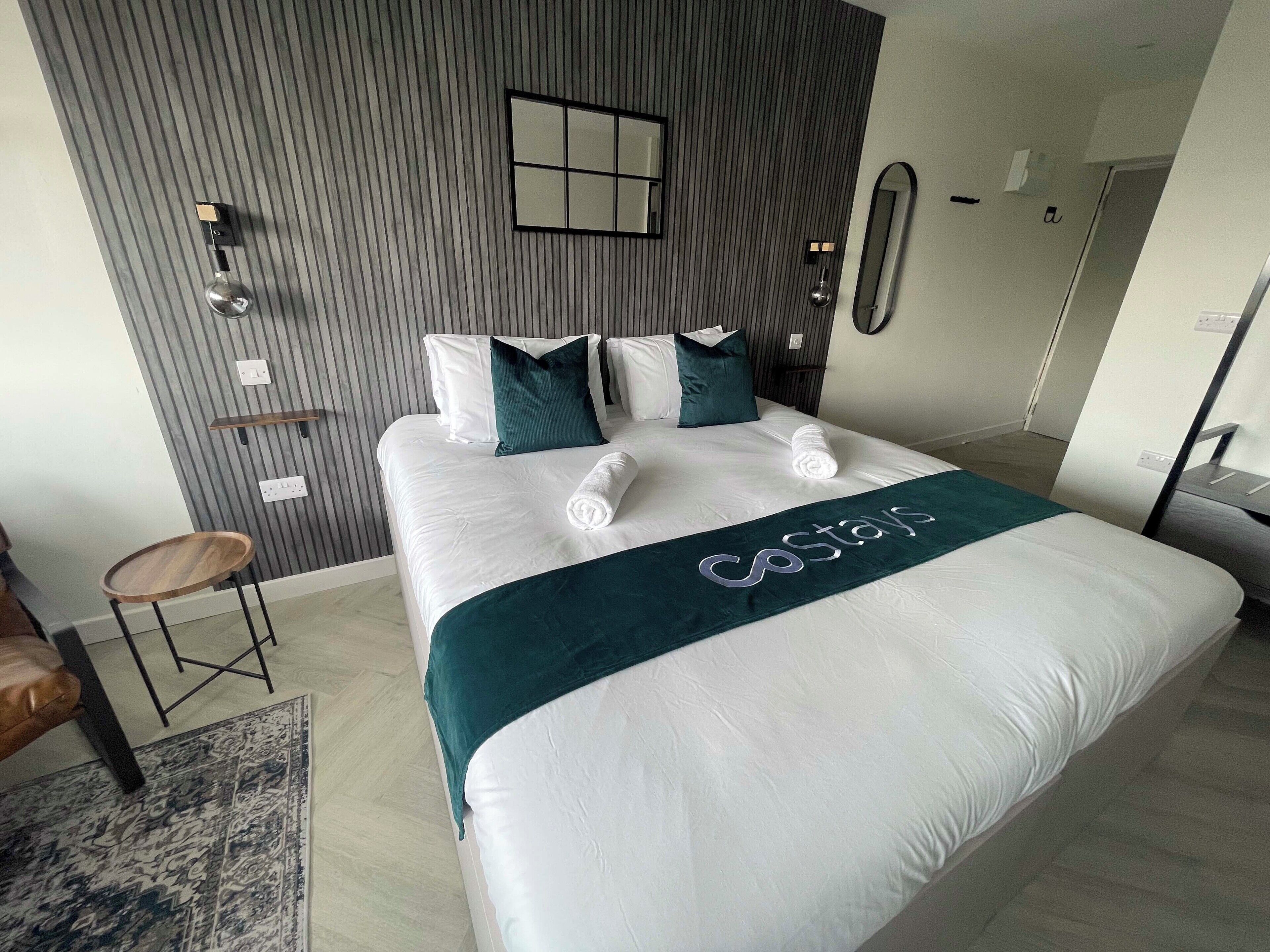 A hotel room with a large bed, a wooden side table, a small stool, a patterned rug, and a modern wall design.