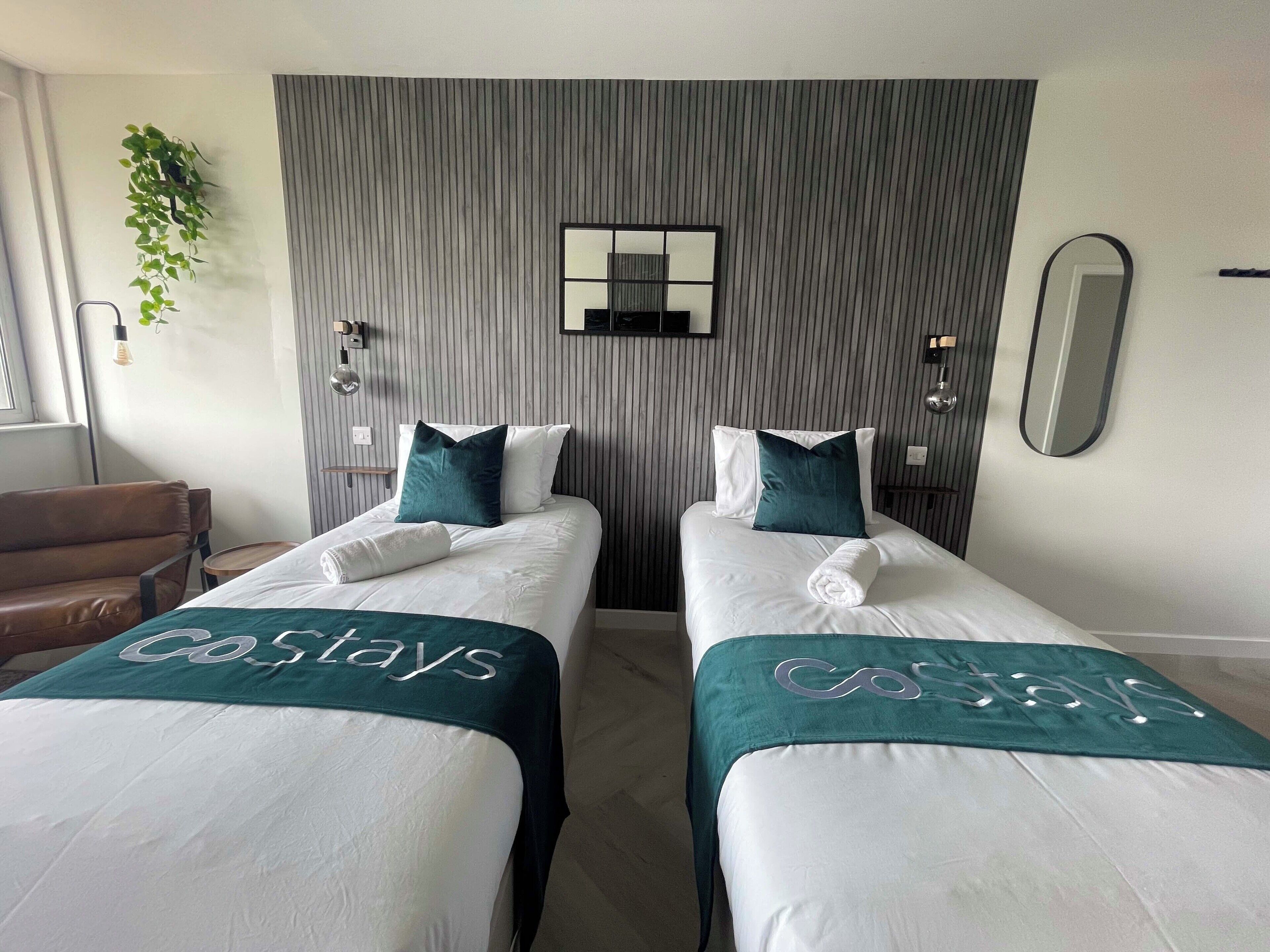A hotel room with two beds, each with a 'Coslays' branded bedspread, a mirror, and a plant on the wall.