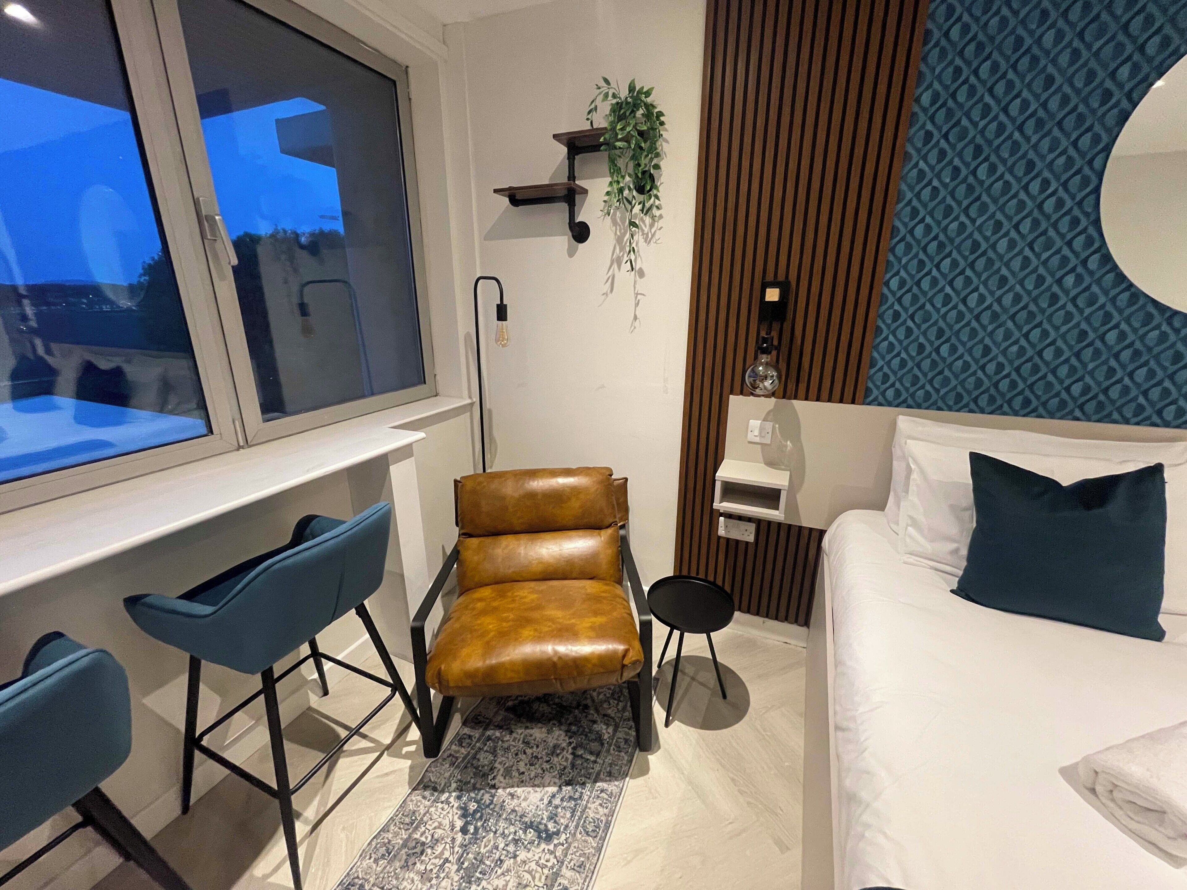 A modern hotel room with a large window, a desk with two chairs, a bed with a pillow, a small plant, and a decorative wall panel.