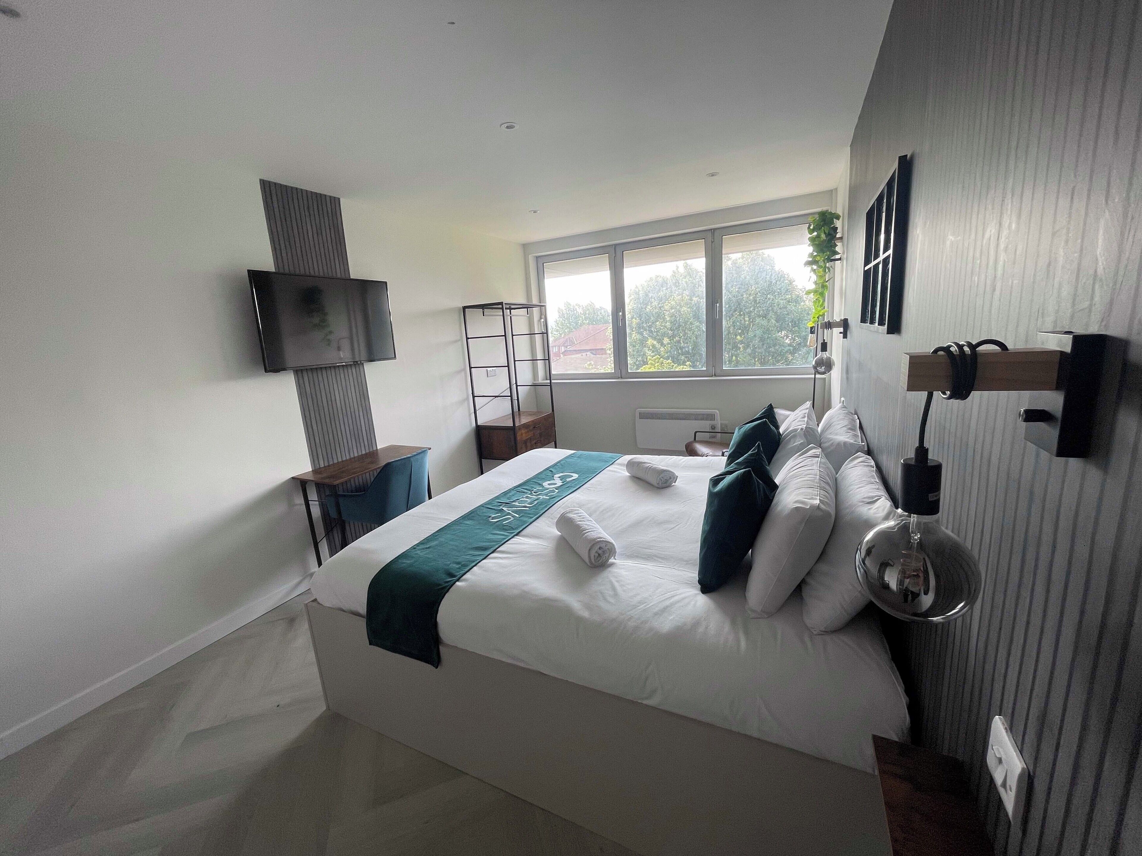 A modern hotel room with a large bed, a desk, a TV, and a window with a view of trees.