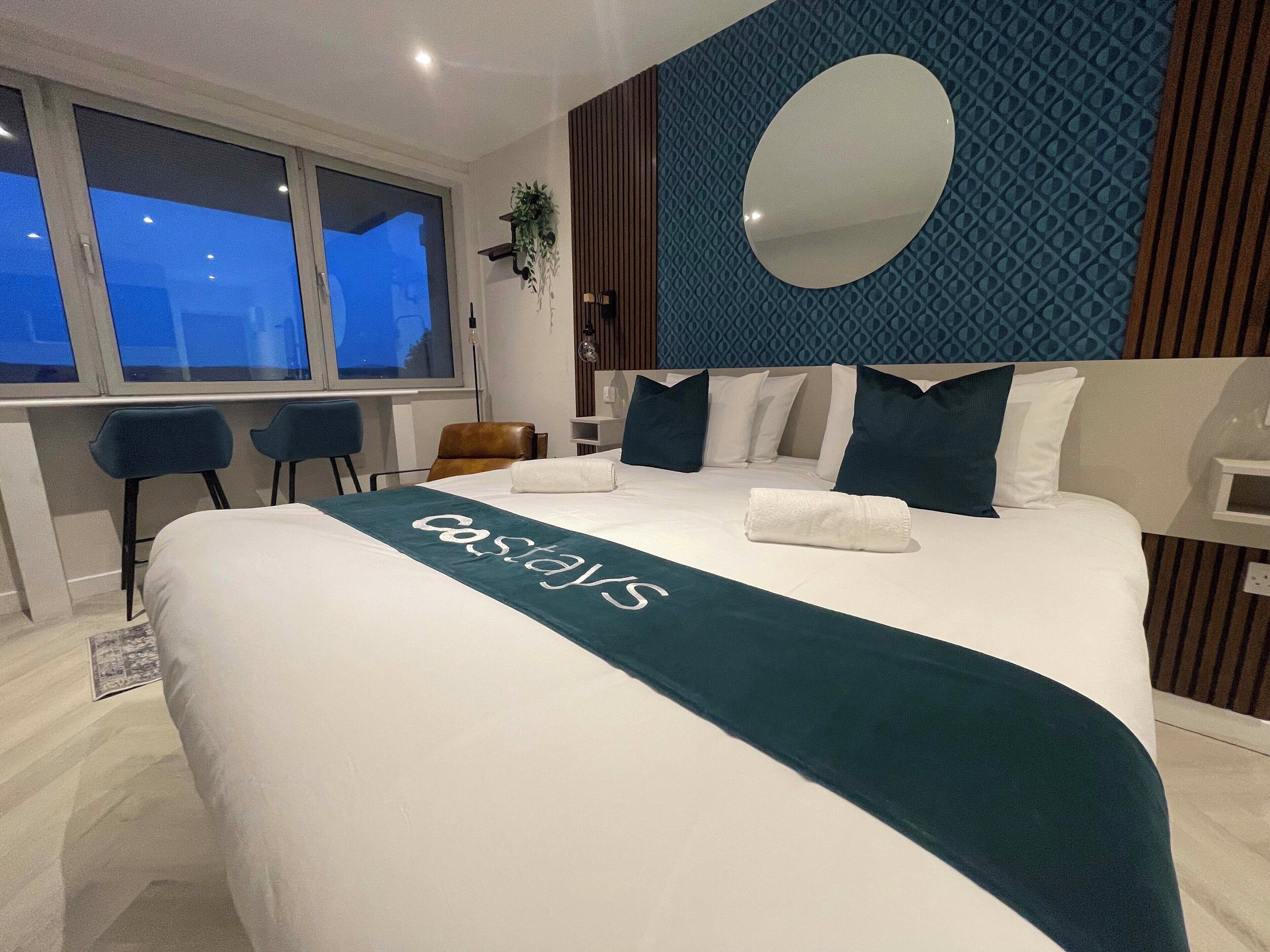 A hotel room with a bed, a desk, two chairs, a large mirror, and a window with a view.