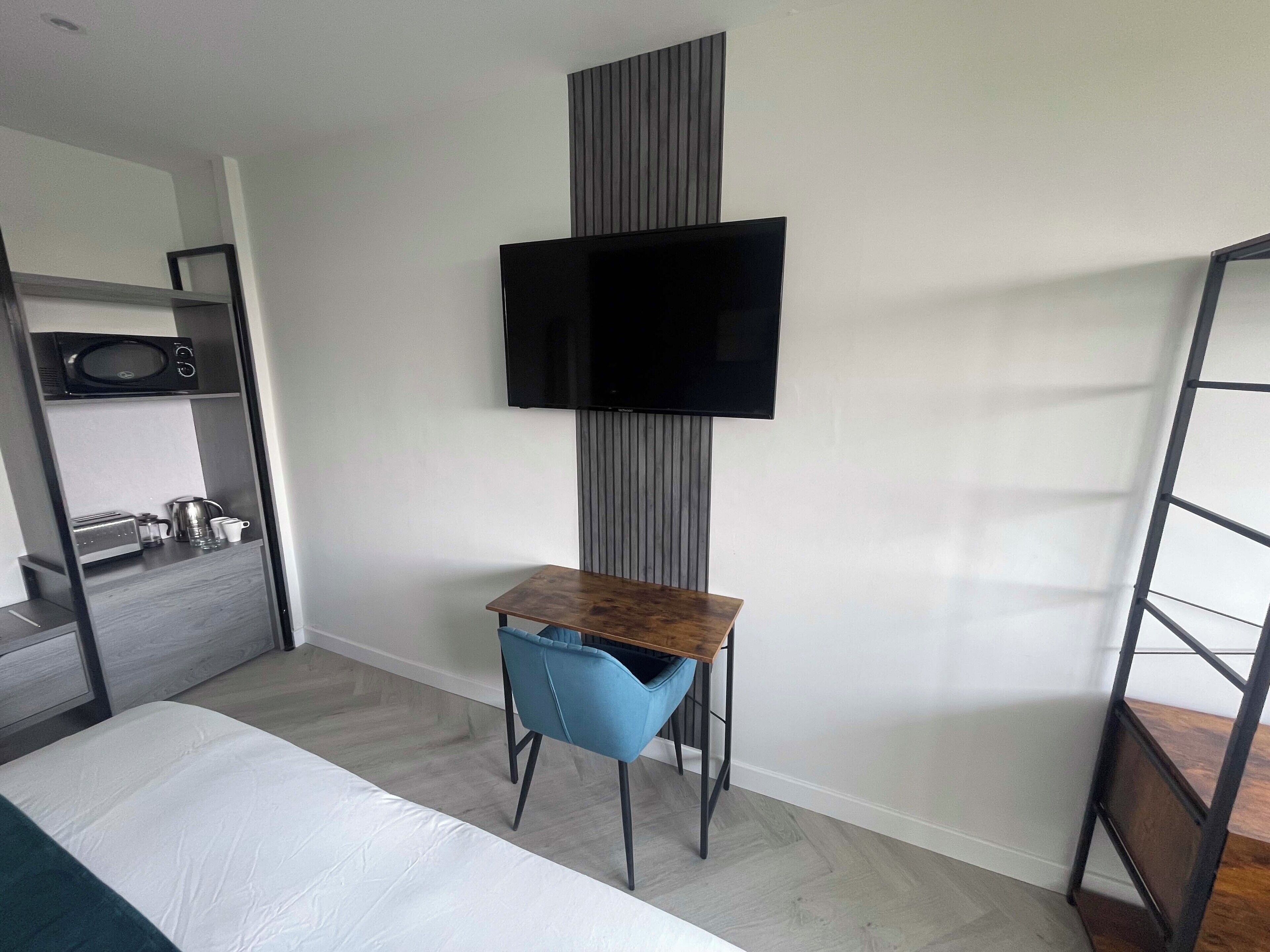 Une chambre d’hôtel moderne équipée d’un lit, d’un bureau avec une chaise, d’un téléviseur à écran plat fixé au mur, d’un four à micro-ondes, d’une machine à café et d’une étagère avec une radio.