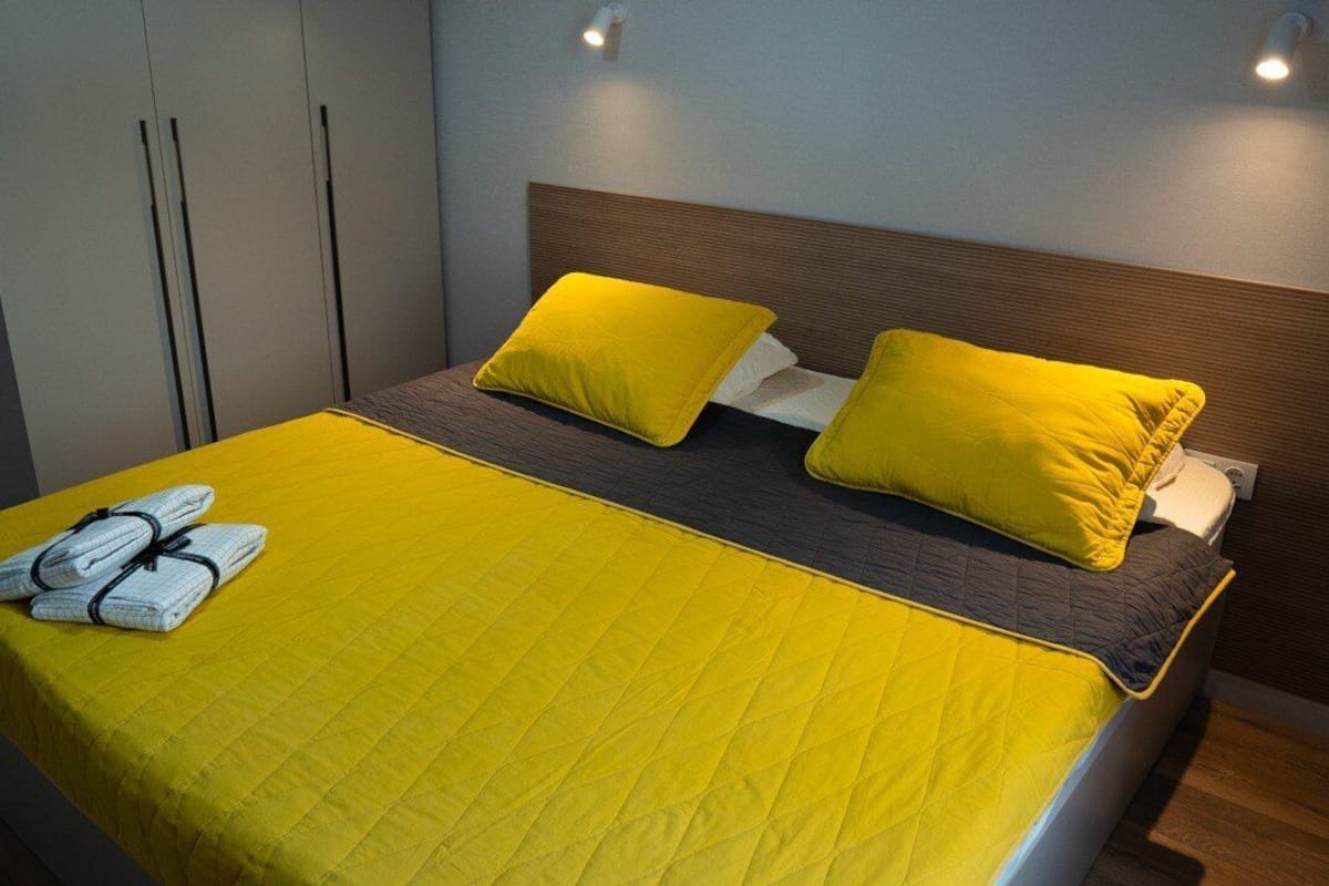 A bed with yellow bedding and pillows, a neatly folded towel, and a wooden headboard.