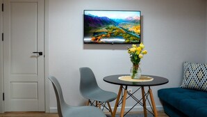 A room with a TV mounted on the wall displaying a scenic landscape, a round table with a vase of yellow flowers, two grey chairs, and a blue sofa. - SingKolSuites (Bishkek)
