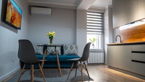 A modern kitchen with a built-in stove, a dining area with a blue sofa and two chairs, and a window with blinds. - SingKolSuites (Bishkek)