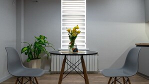 A room with a round table, two grey chairs, a potted plant, a vase with yellow flowers, and a window with white blinds. - SingKolSuites (Bishkek)