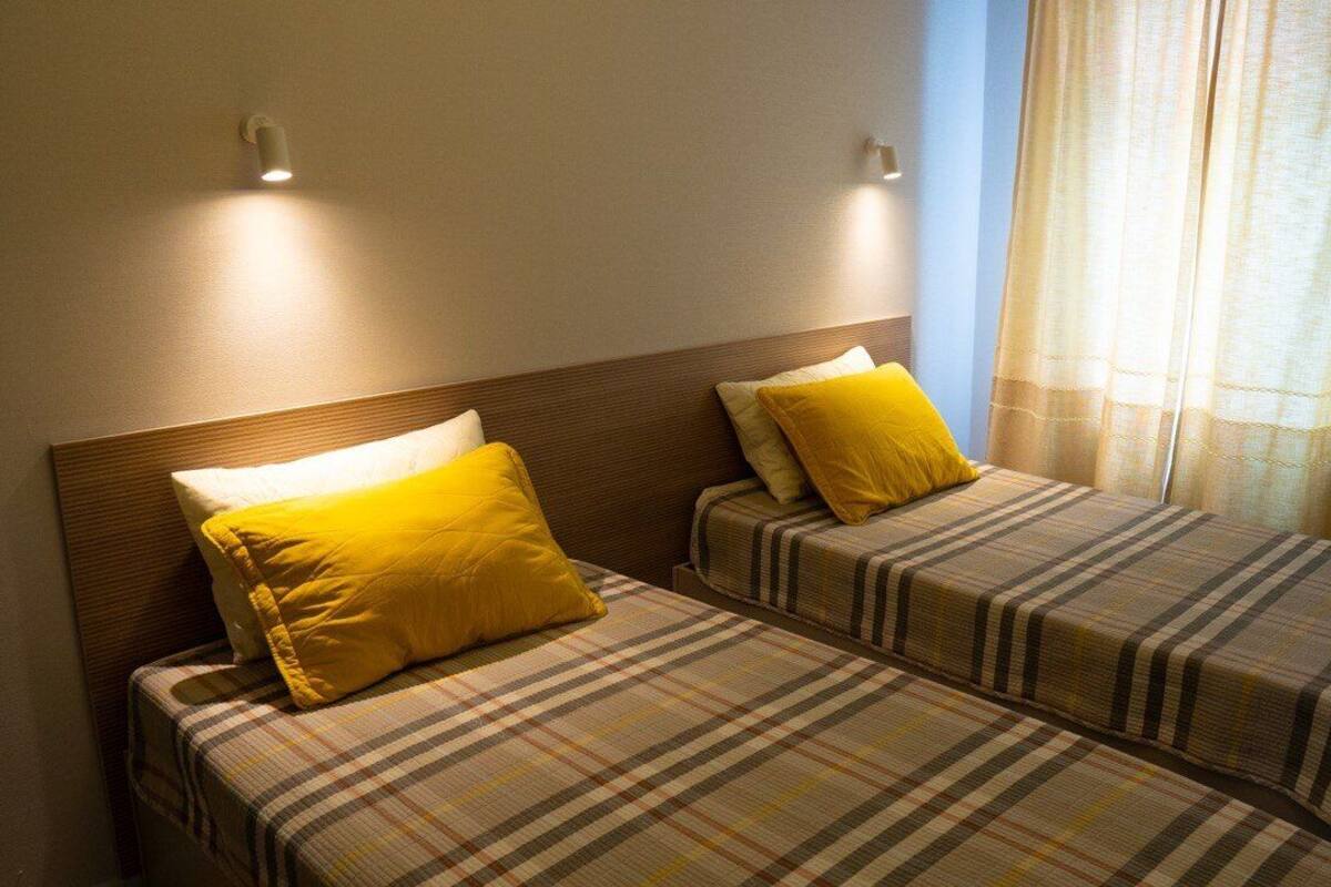 A room with two beds, each with a plaid bedspread and yellow pillows. There are wall-mounted lights above the beds and a window with curtains.