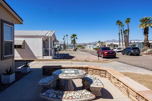 Un barrio residencial con camino pavimentado, un patio con mesa y bancos de mosaico, y varios coches aparcados.