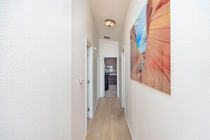 Un couloir étroit, éclairé par un luminaire au plafond, avec une porte donnant sur une pièce et un grand tableau accroché au mur.