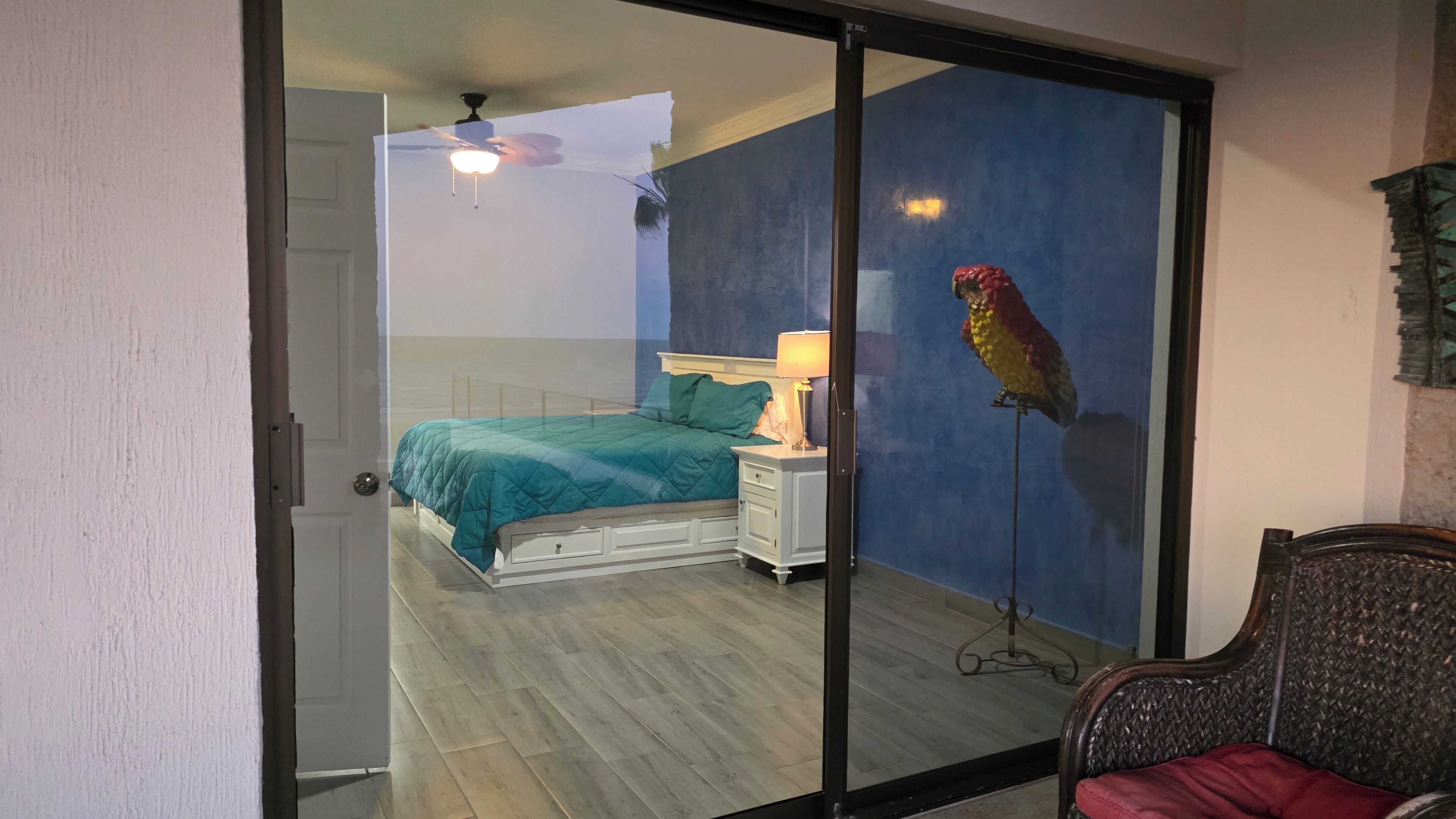Een slaapkamer met een bed, nachtkastje, lamp en een papegaai op een standaard.