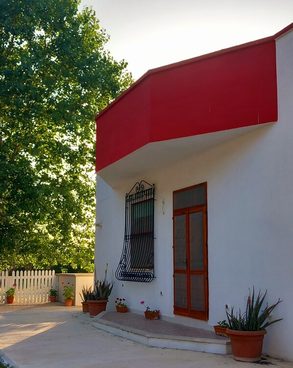Un edificio de fachada blanca y techo rojo, con una ventana protegida por una reja metálica. Hay macetas con plantas en el suelo frente al edificio.