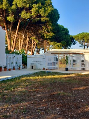 Un edificio blanco con un jardín cercado, plantas en macetas y un árbol grande al fondo.