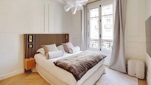 A bedroom with a large bed, a wooden headboard, a bedside table, a nightstand, a window with curtains, and a view of buildings outside. - Elegant A/C Apt 2BR/6P – Paris 17th/Monceau (Paris)