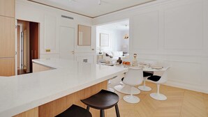 A modern kitchen with a white countertop, wooden cabinets, and a dining area with white chairs and a table. - Elegant A/C Apt 2BR/6P – Paris 17th/Monceau (Paris)
