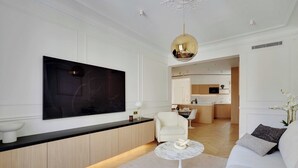 A modern living room with a large flat-screen TV, a white sofa, a round coffee table, and a minimalist kitchen in the background. - Elegant A/C Apt 2BR/6P – Paris 17th/Monceau (Paris)