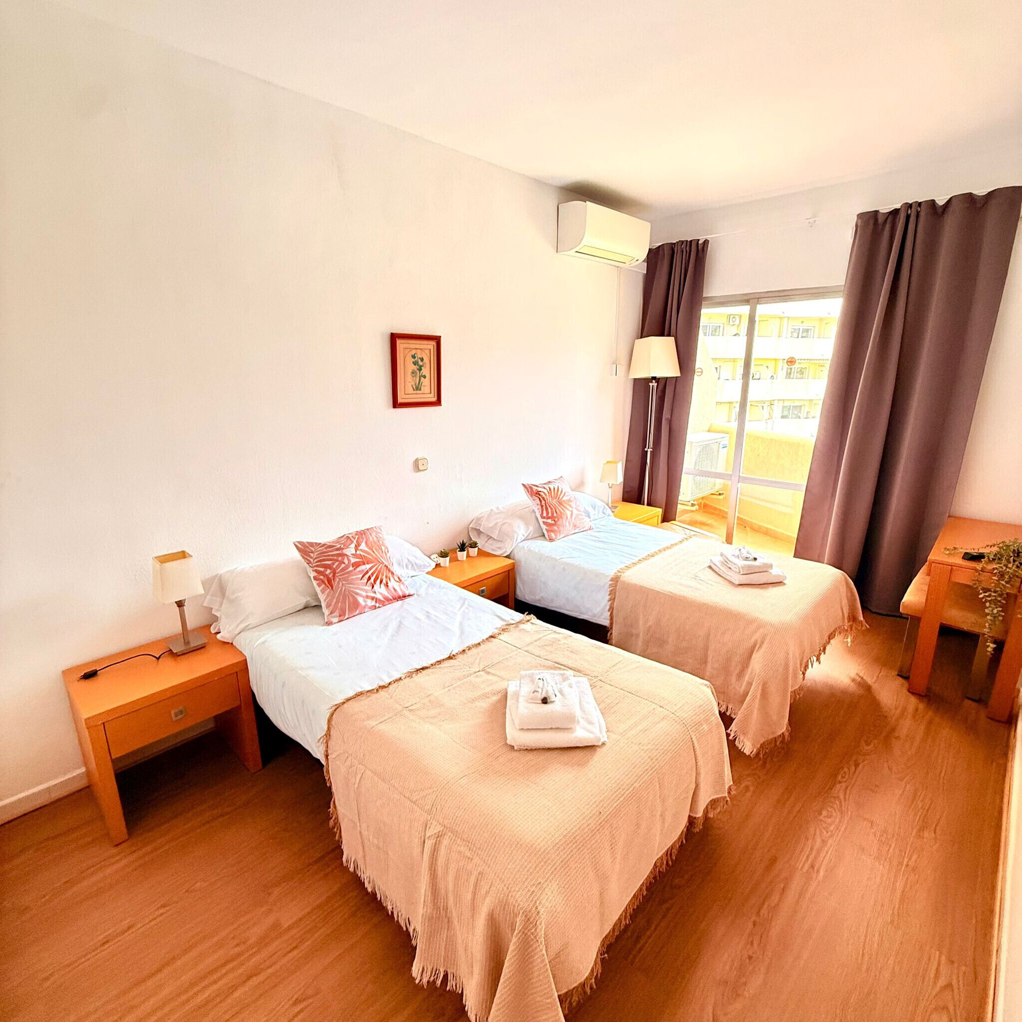 A hotel room with two beds, a bedside table with a lamp, a small plant, and a window with curtains.