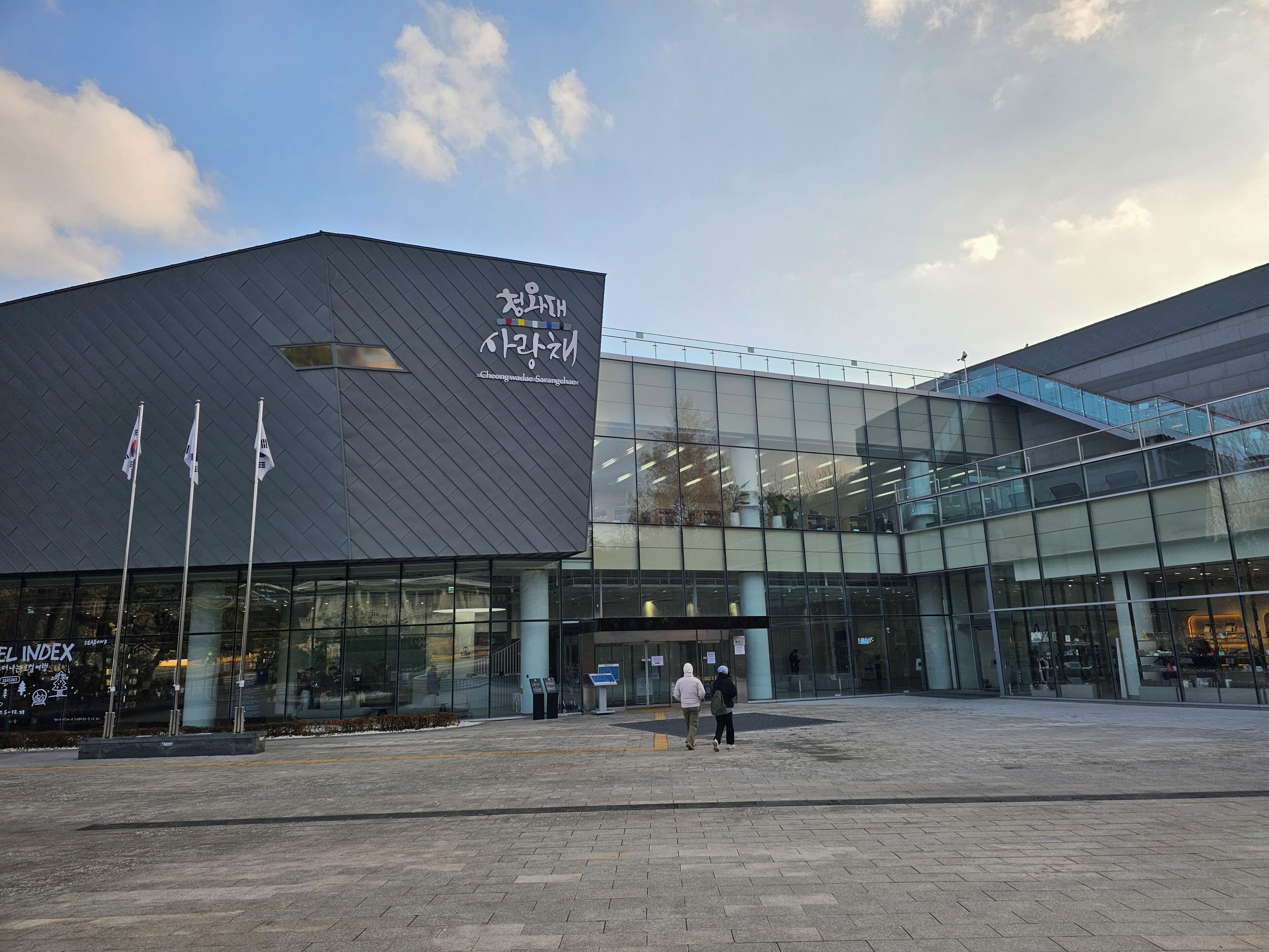A modern building with a dark facade and large glass windows. There are three flags in front of the building.