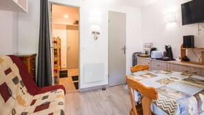 A compact room with a bed, a small table, a chair, a TV, and a kitchenette.