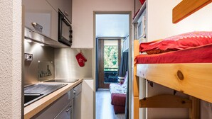 A compact kitchen with a sink and cabinets, a wooden bed with red bedding, and a view of an outdoor area through an open door.