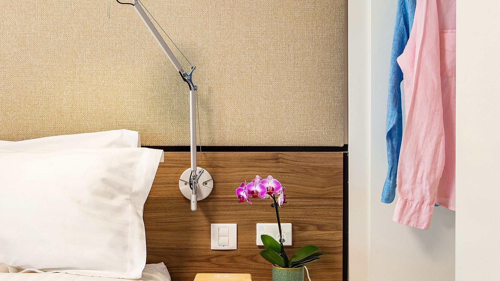 A neatly made bed with a white pillow, a bedside lamp, a small potted orchid, and a wall-mounted light switch.