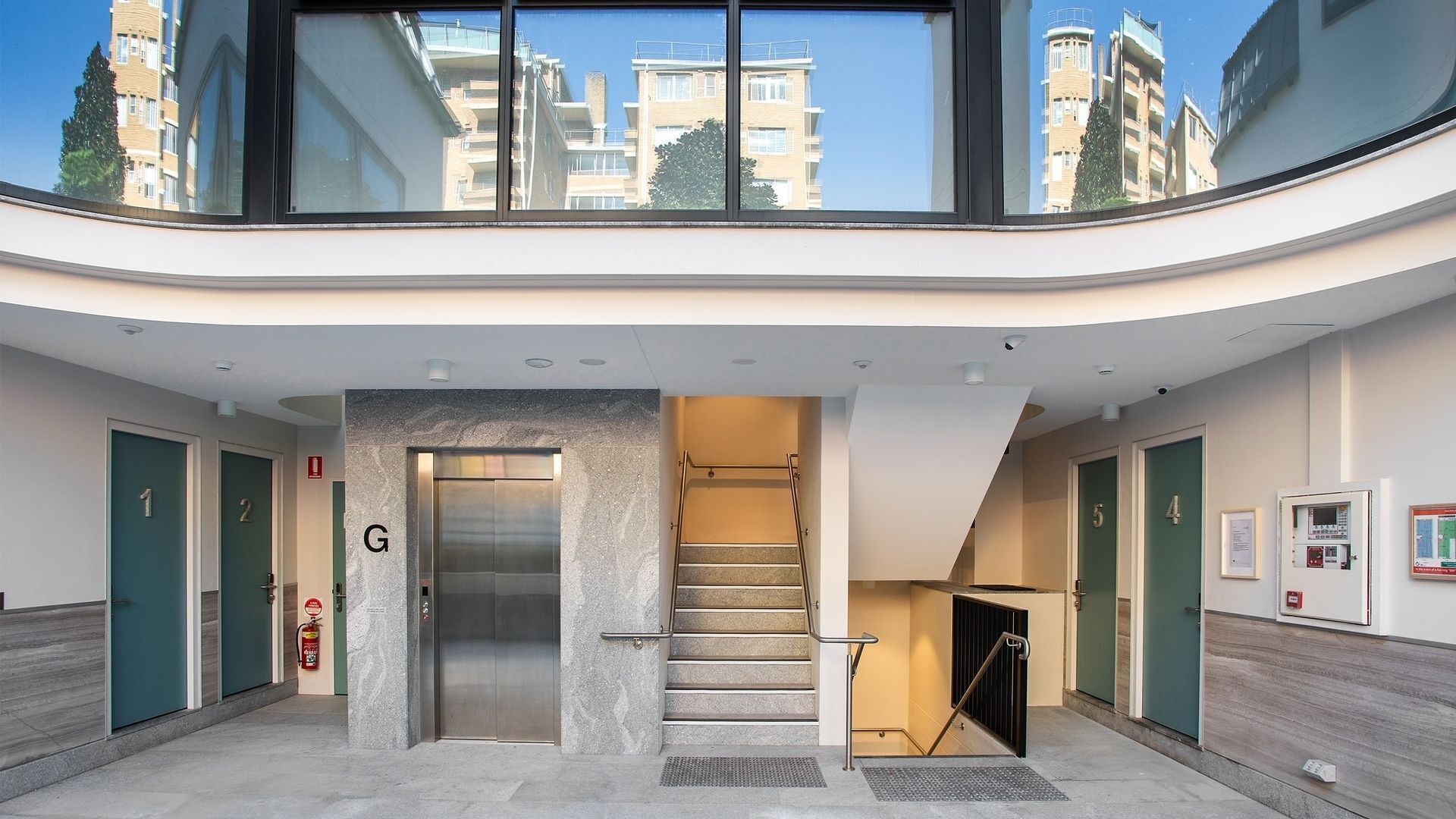 A hotel lobby with a central elevator, a staircase, and a view of the surrounding buildings.