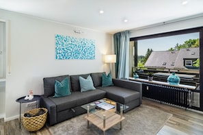 Ein modernes Wohnzimmer mit einem grauen Ecksofa, einem Glastisch und Blick auf ein Haus durch ein großes Fenster.