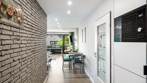 Un pasillo estrecho con pared de ladrillo, un comedor con sillas y una mesa, y una cocina con armarios y un reloj.