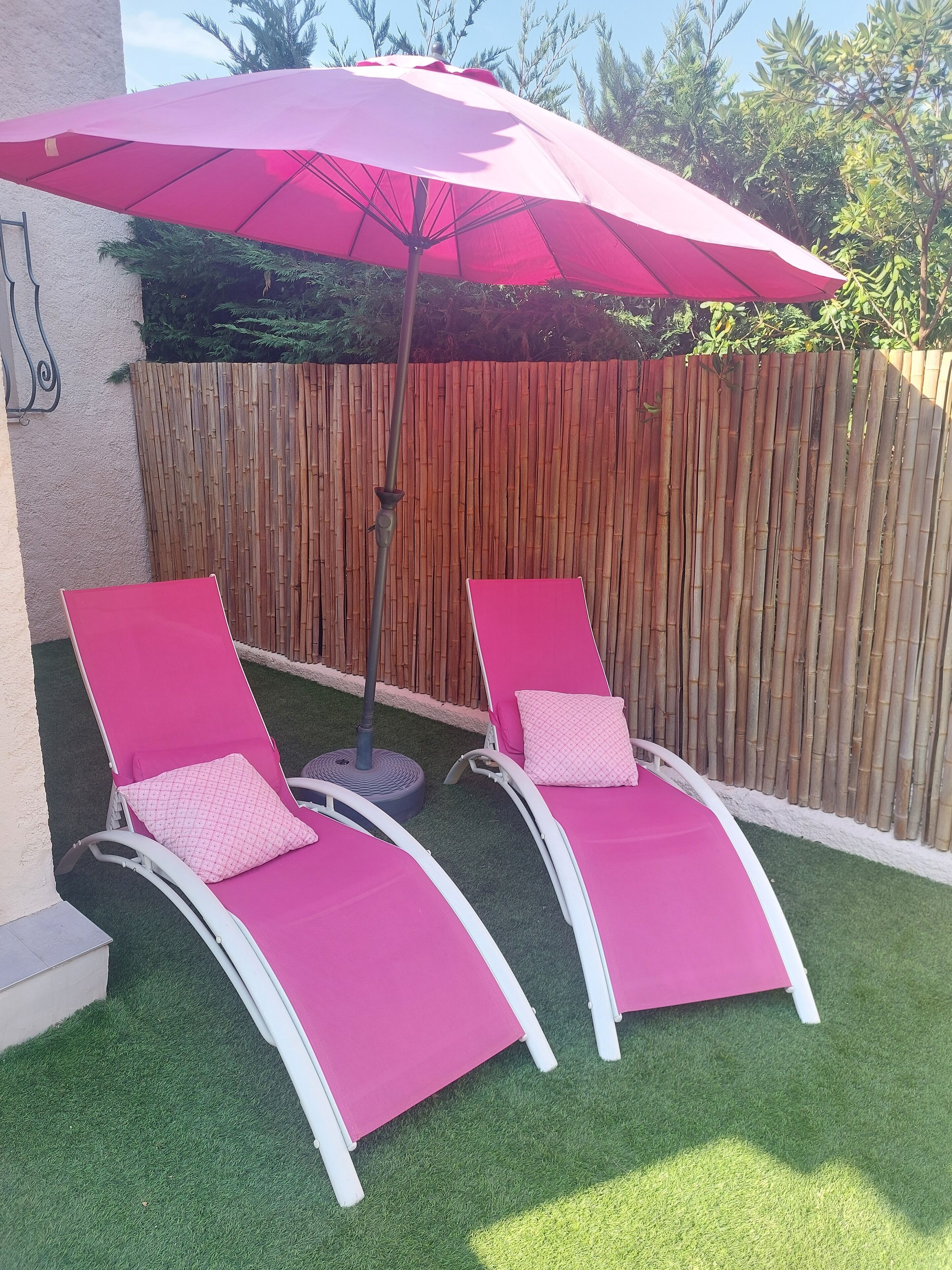 Two pink lounge chairs with pillows, a red umbrella, and a bamboo fence in a backyard.