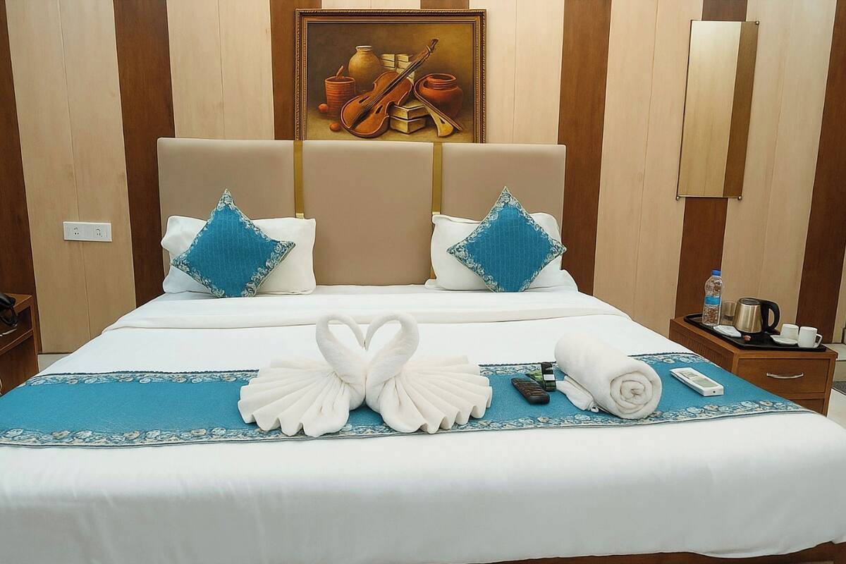 Cama bem arrumada com edredom azul e branco, duas almofadas, uma toalha e um arranjo de toalhas em formato de cisne. Um quadro está pendurado acima da cama, e uma mesinha de cabeceira com chaleira, xícaras e garrafa são visíveis.