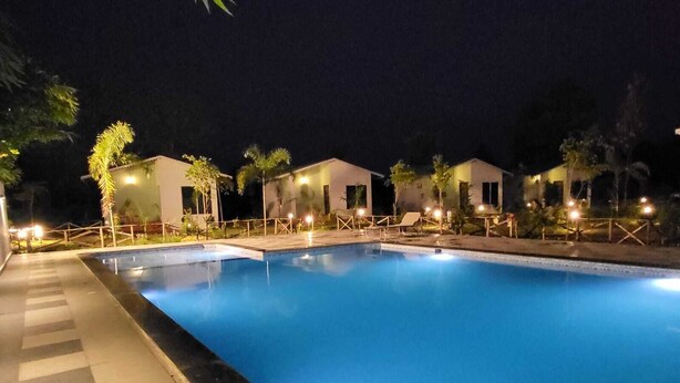 A swimming pool with adjacent lounge areas, surrounded by small buildings with outdoor lighting. - Pench Kulamama Safari Resort (Kurai)