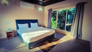A bedroom with a bed, a nightstand, a window with curtains, and a view of greenery outside. - Pench Kulamama Safari Resort (Kurai)