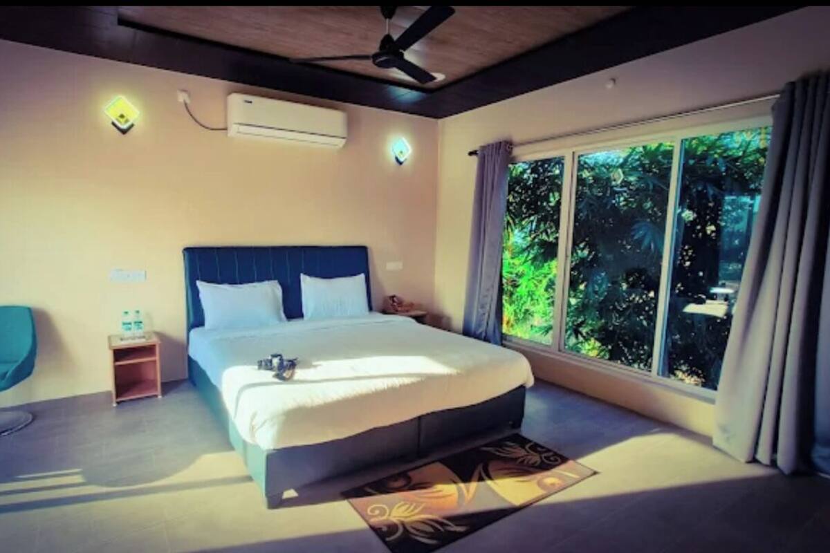 Ein modernes Hotelzimmer mit einem großen Bett, einem blauen Sessel, einem kleinen Tisch und einem Fenster mit Blick auf Grünflächen.