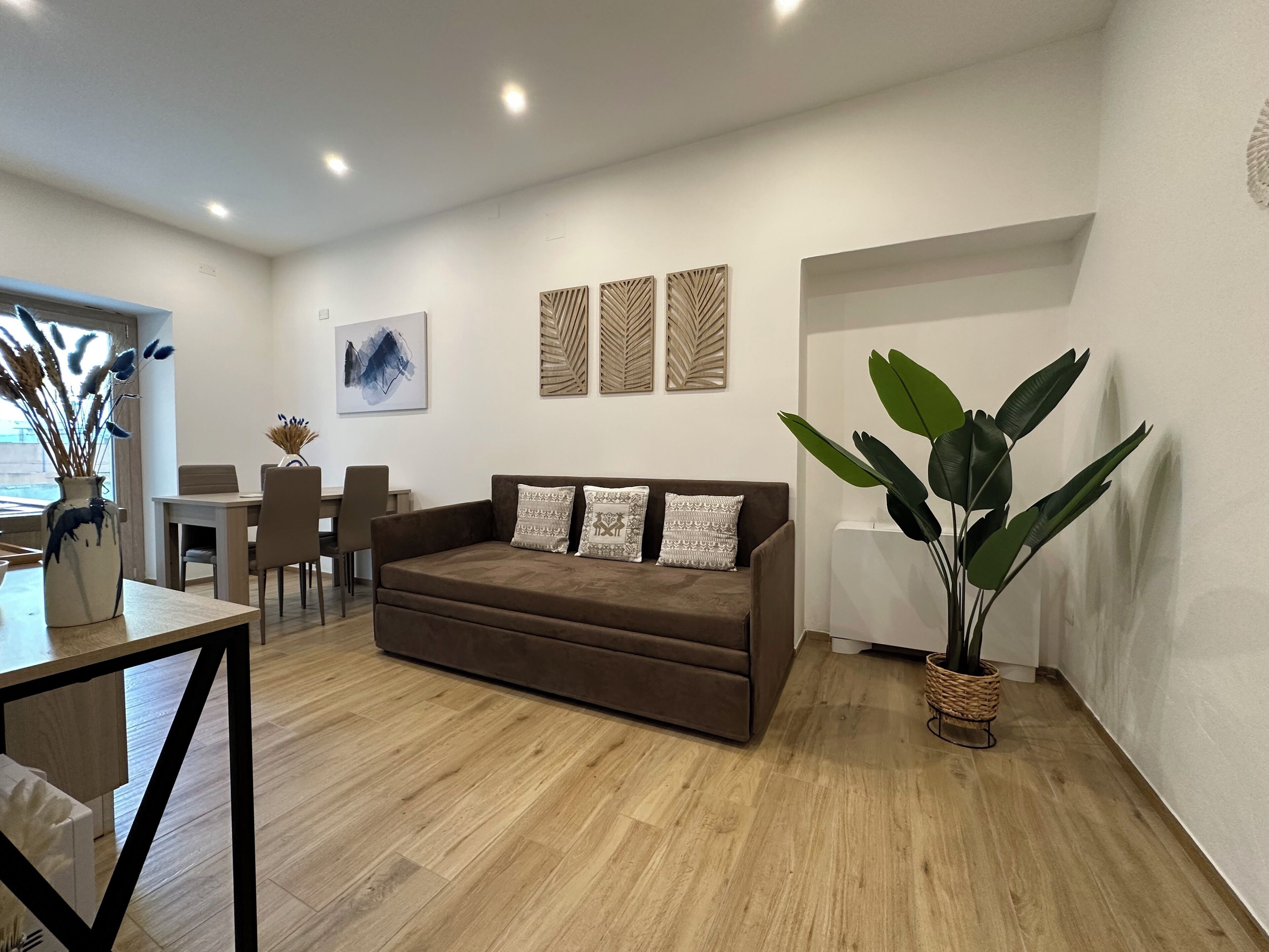 Una sala de estar moderna con un sofá marrón, una mesa de comedor y sillas. Se observan paneles decorativos en la pared y una planta grande.