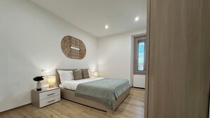 A bedroom with a bed, bedside tables, a wall-mounted circular light, a window with blinds, and a wooden floor. - NG Collection il Barbagia (Alghero)