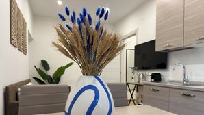 A modern kitchen with wooden cabinets, a dining table with a vase of wheat and blue flowers, and a sofa in the background. - NG Collection il Barbagia (Alghero)