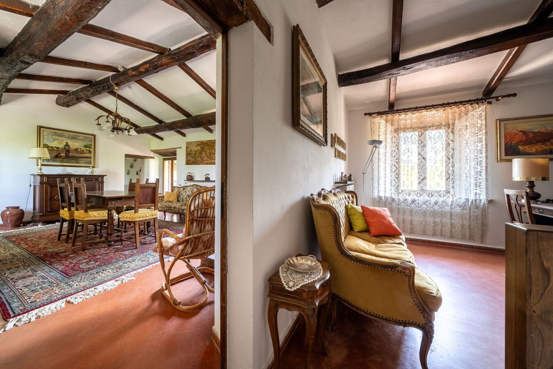 A traditional room with exposed wooden beams, a dining area with a table and chairs, a rocking chair, and a window with lace curtains.
