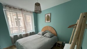 Quarto com cama, mesa de cabeceira pequena, luminária pendente e janela com cortinas leves.