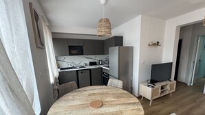 Cozinha moderna com forno embutido, micro-ondas e frigorífico. Há uma mesa de jantar com cadeiras e uma televisão sobre um móvel.