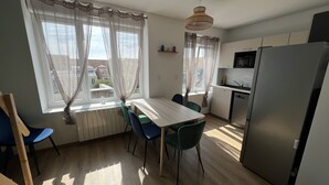 A compact kitchen with a dining table and chairs, a refrigerator, and a window with sheer curtains.