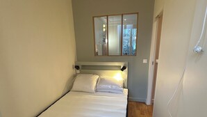 Una habitación pequeña con una cama individual, suelo de madera, cabecera con iluminación integrada, un espejo sobre la cama y una puerta que da a otra habitación.