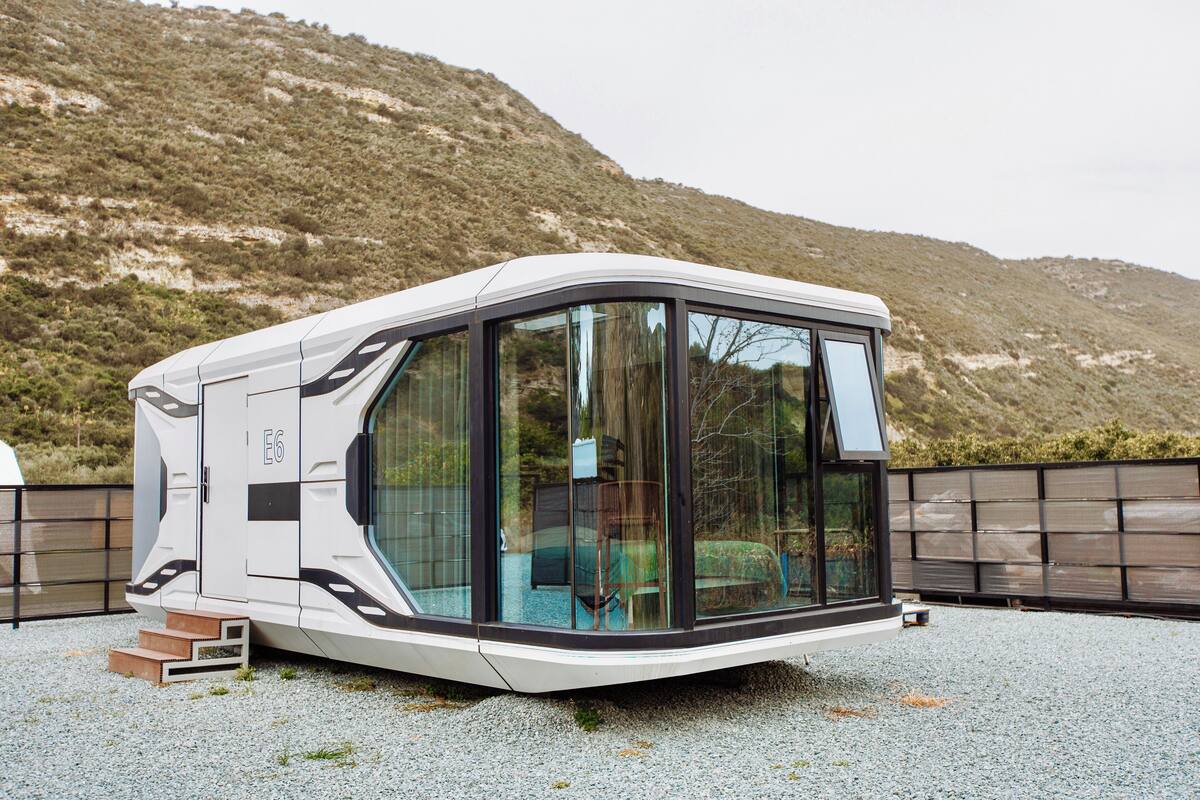 A modern, compact RV with a curved design, large windows, and a flat roof, situated on a gravel surface with a hilly backdrop.