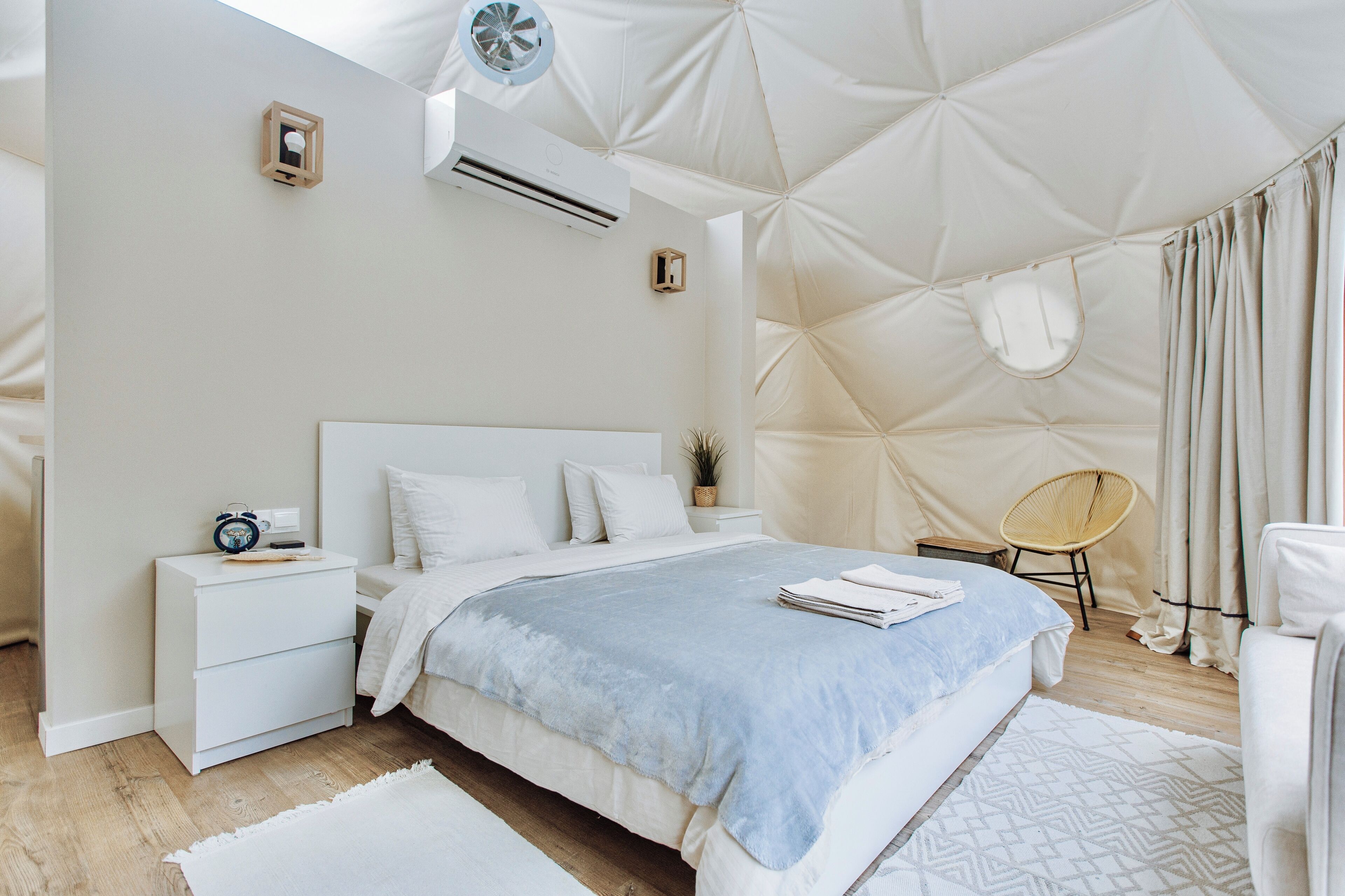 Een moderne slaapkamer met een groot bed, witte nachtkastjes, een klein tafeltje, een stoel en een uniek geometrisch plafondontwerp.