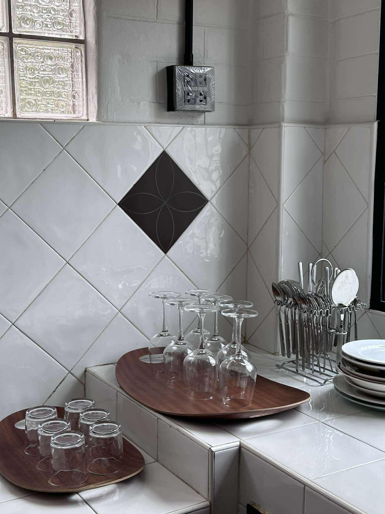 A kitchen with white tiles, a black decorative tile, a power outlet, a wine rack, and a tray with glasses.