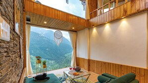 Ein gemütliches Zimmer mit einem grünen Sofa, einem kleinen Tisch mit einer Dekoration und Blick auf eine gebirgige Landschaft durch ein großes Fenster.