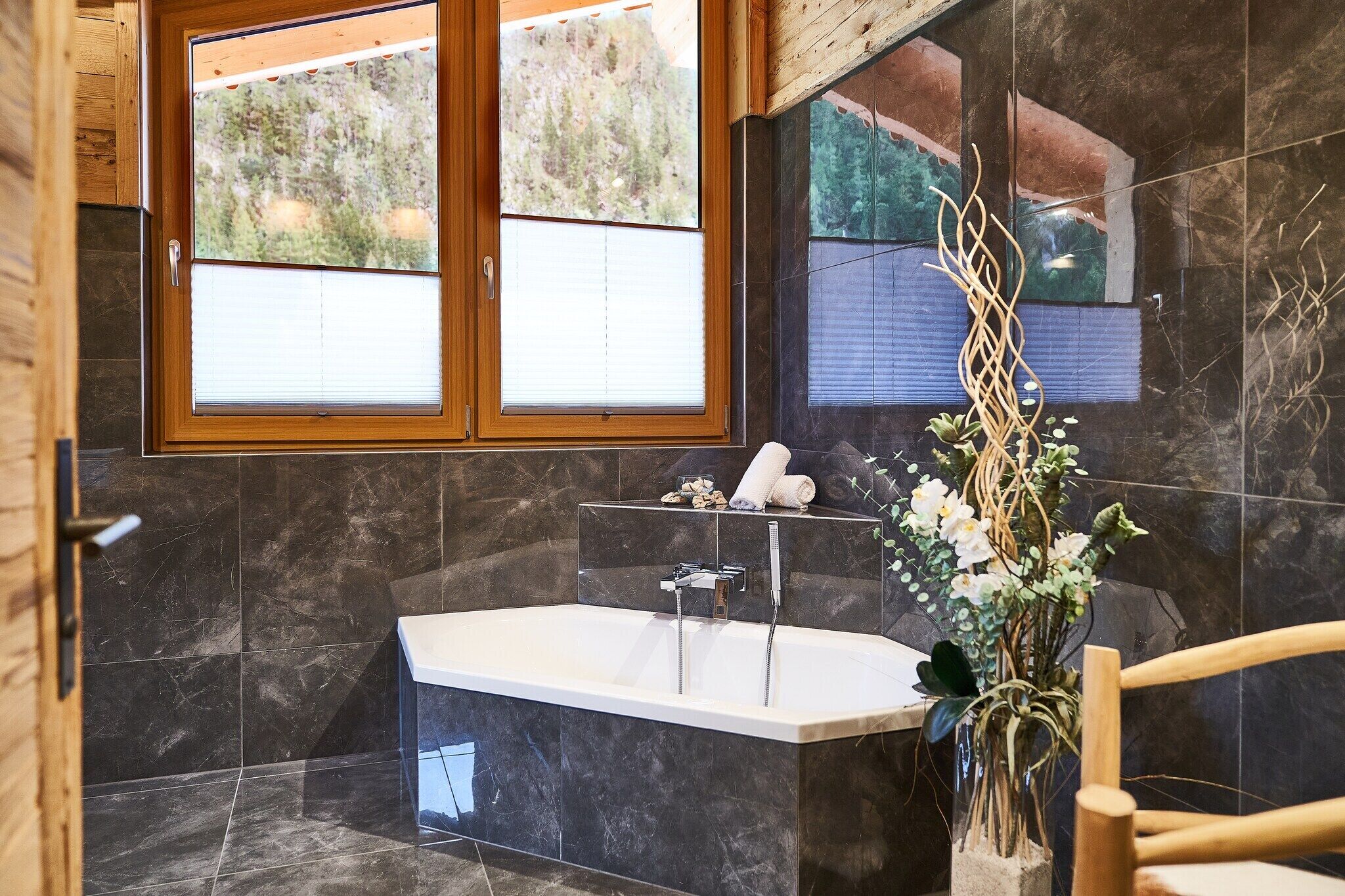 A modern bathroom with a freestanding bathtub, dark marble tiles, a wooden-framed window, and a decorative plant.