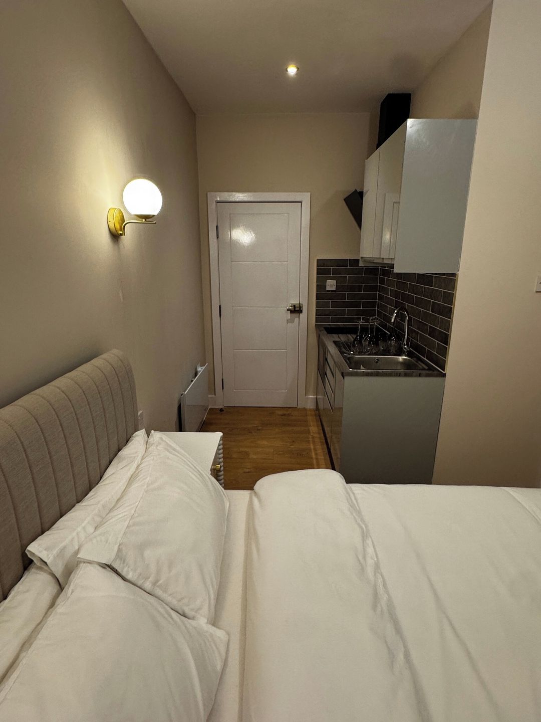 A compact room with a bed, a bedside table, a wall-mounted light, a door, and a kitchenette area with a sink and cabinets.