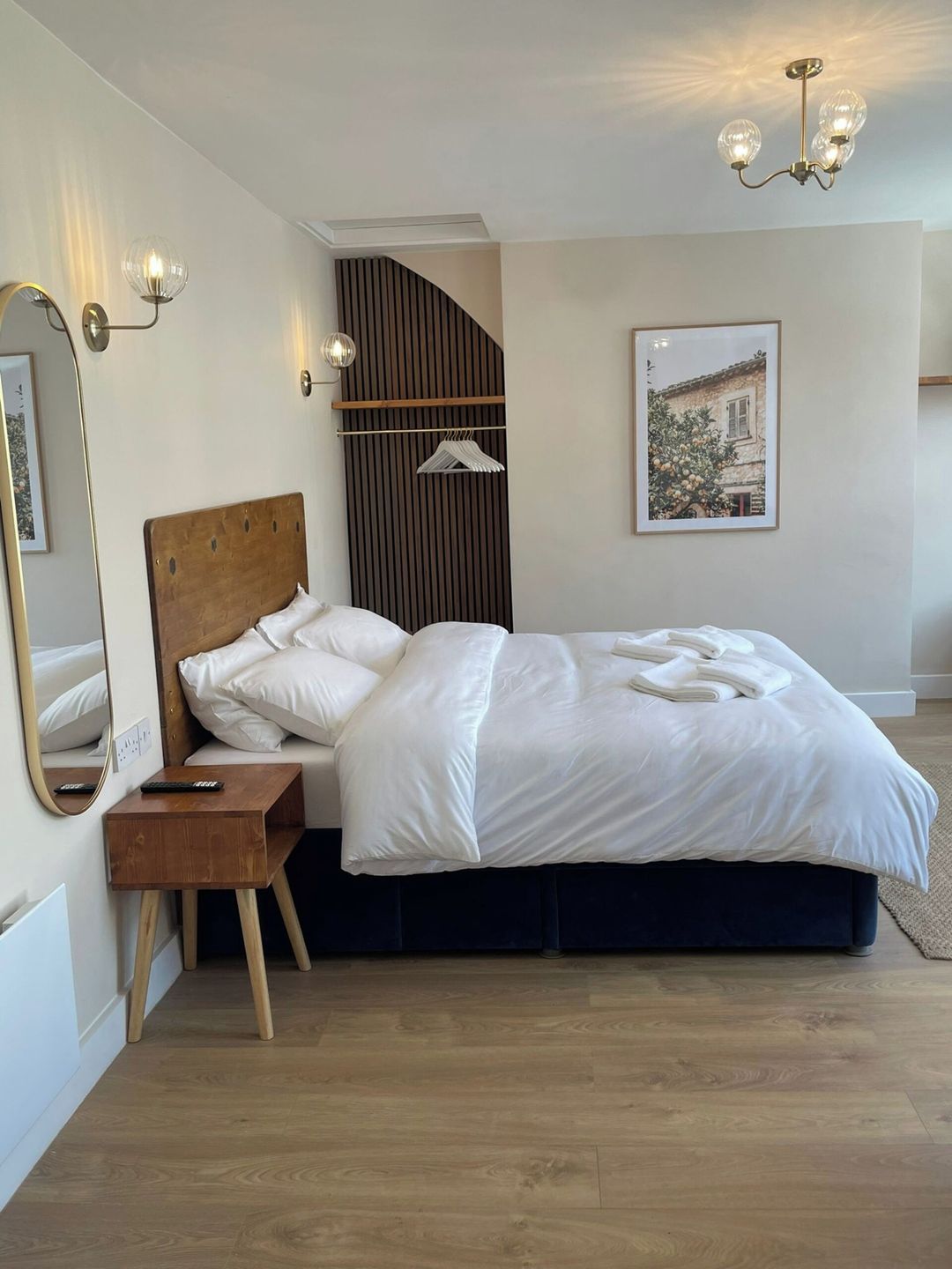 A bedroom with a wooden headboard, a bed with white linens, a bedside table with a remote control, a hanging light fixture, and a framed picture on the wall.